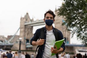 Portrait of a happy student wearing a facemask on the street in London and holding a notebook while looking at the camera - pandemic lifestyle concepts